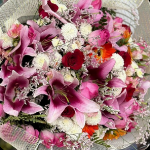Buquê com lírios rosa, rosas vermelhas, gérberas laranjas e flores brancas decorativas.